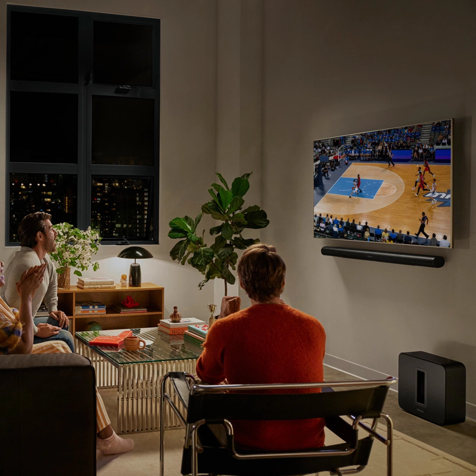 Lifestyle image of two males and a female in a living room enjoying watching basketball on a wall mounted TV with the Sonos Arc Ultra Soundbar Black wall mounted underneath and Sonos Sub 4 black on the floor.