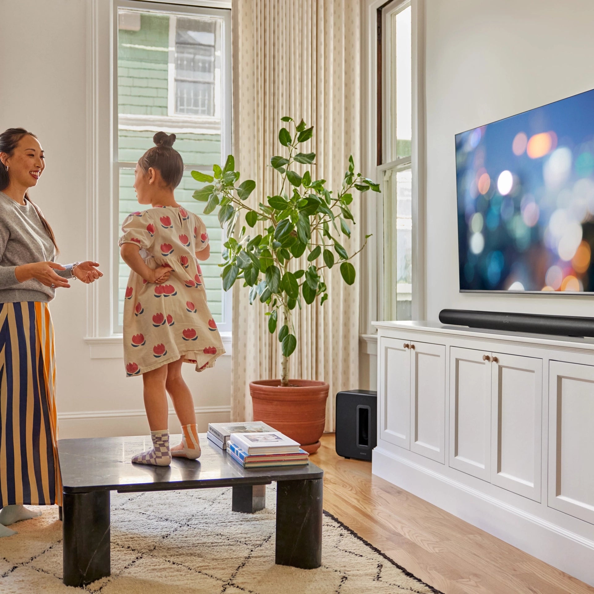 Lifestyle image of a woman and child playing in a living room with the TV on next to them and the Sonos Arc Ultra Black and Sonos Sub 4 Black in view.