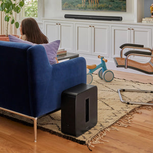Lifestyle image of a living room with a female watching TV which is wall mounted and Sonos Arc Ultra Black on a stand underneath, there is a Sonos Sub 4 Subwoofer Black next to the sofa the female is sitting on.